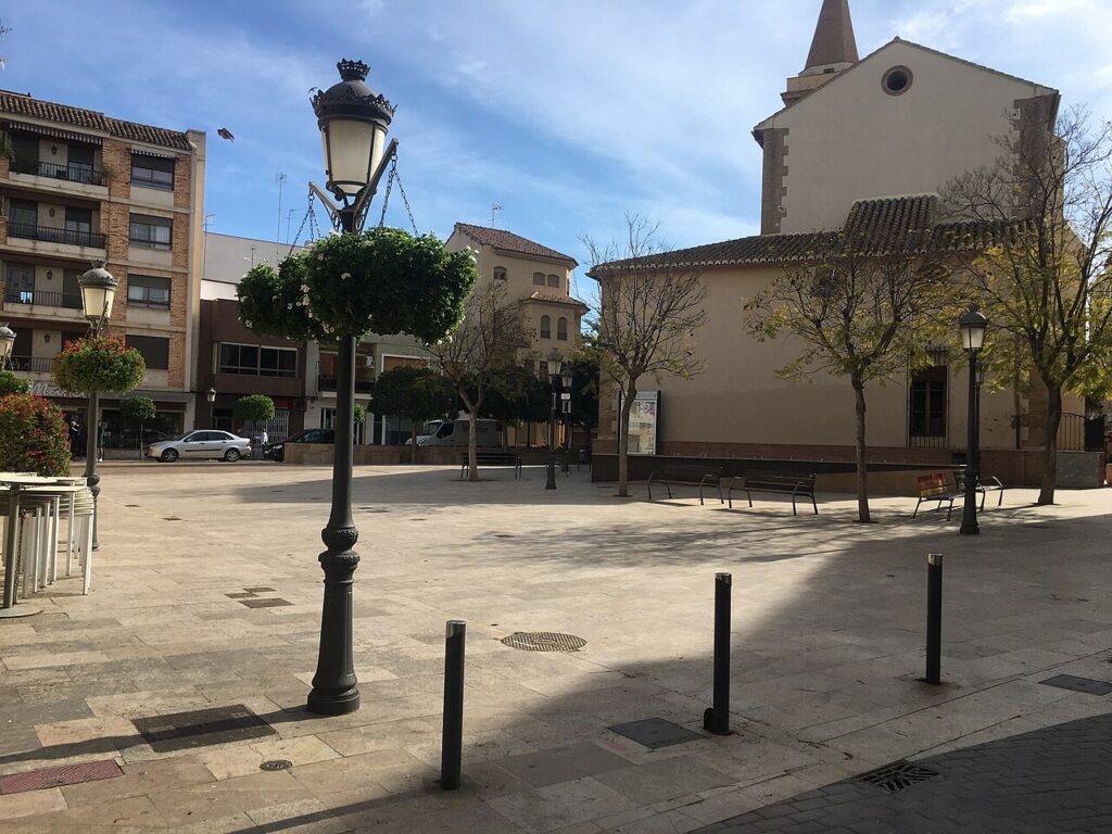 The sunny Plaça del País Valencià in L'Eliana with a church tower, street lamps and apartment buildings