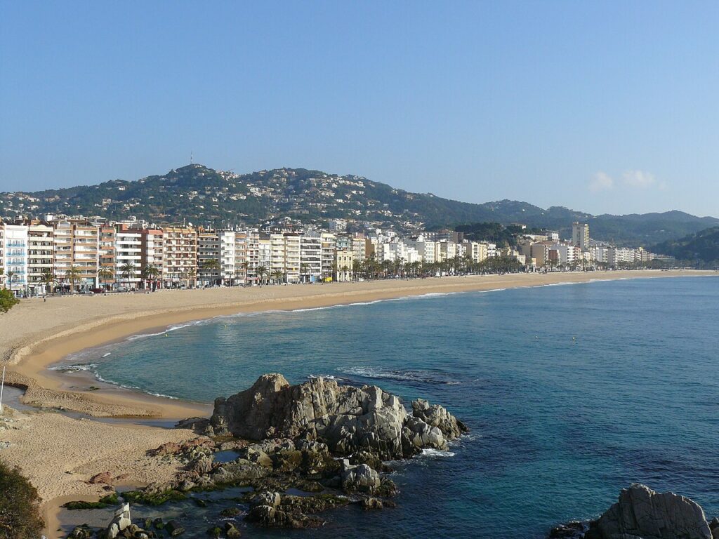 The main beach of Lloret de Mar with rocks in the blue sea and the seafront apartment buildings below green hills