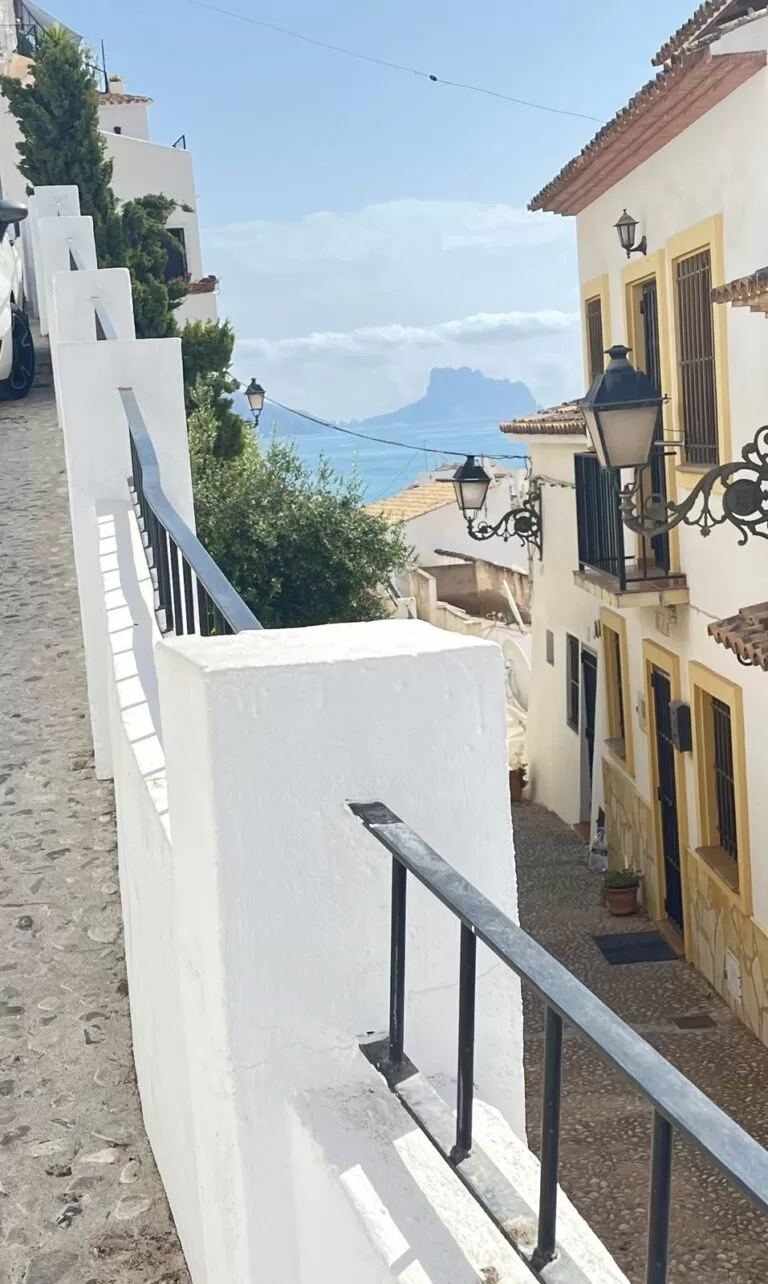 A steep whitewashed stepped street in Altea old town with steel handrails, looking out to the sea and the distant Peñón de Ifach