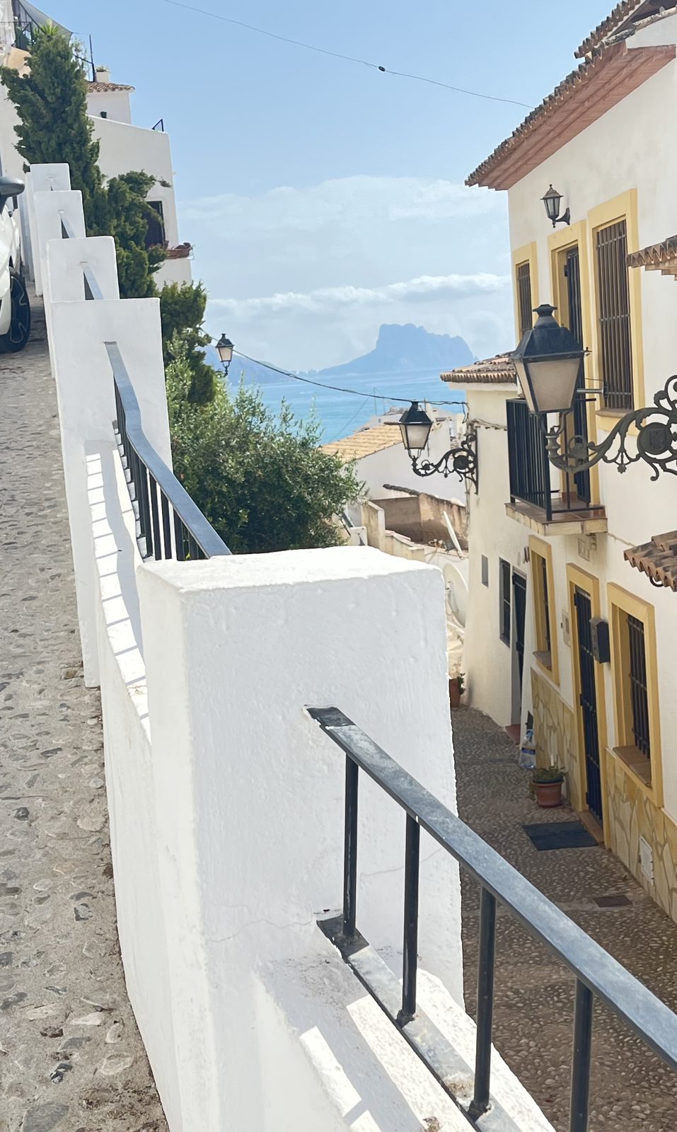 A steep whitewashed stepped street in Altea old town with steel handrails, looking out to the sea and the distant Peñón de Ifach