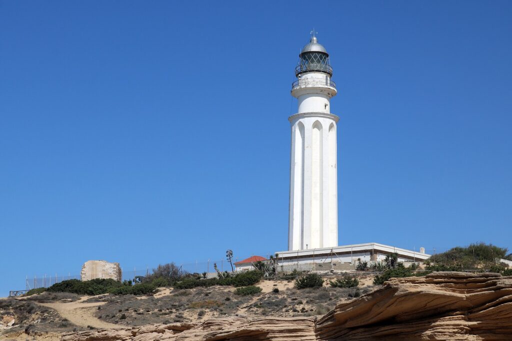 The white Cabo de Trafalgar lighthouse and a ruined stone tower on sandy dunes above rocky ledges.