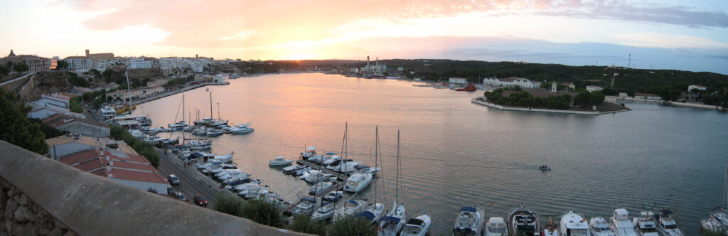 Elevated panoramic view along the long natural harbour of Mao in Menorca at sunset, with moored yachts and motor boats in the marina below and wooded shores stretching towards the sea.