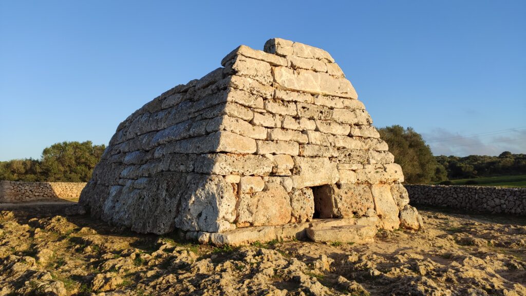 The Naveta des Tudons, a boat-shaped prehistoric burial monument built of stacked dry-stone blocks, standing on rocky ground in low golden sunlight under a clear blue sky.