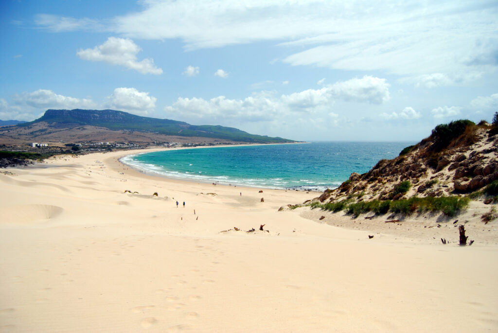 View from the top of the great sand dune at Bolonia looking down over a long curving white beach, turquoise sea and green hills under a partly cloudy sky.