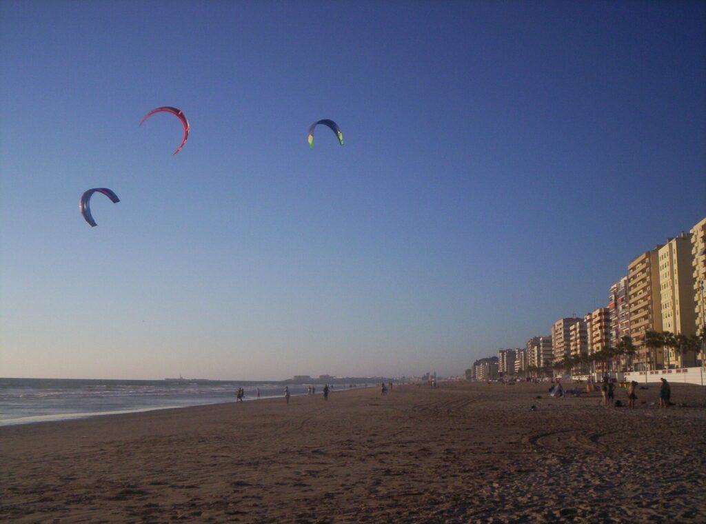 Wide sandy Playa de la Victoria in Cadiz with kites overhead and apartment blocks along the seafront.