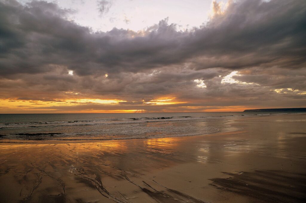 Broad flat beach at low tide under a dramatic cloudy sky at Zahara de los Atunes, with hills beyond.