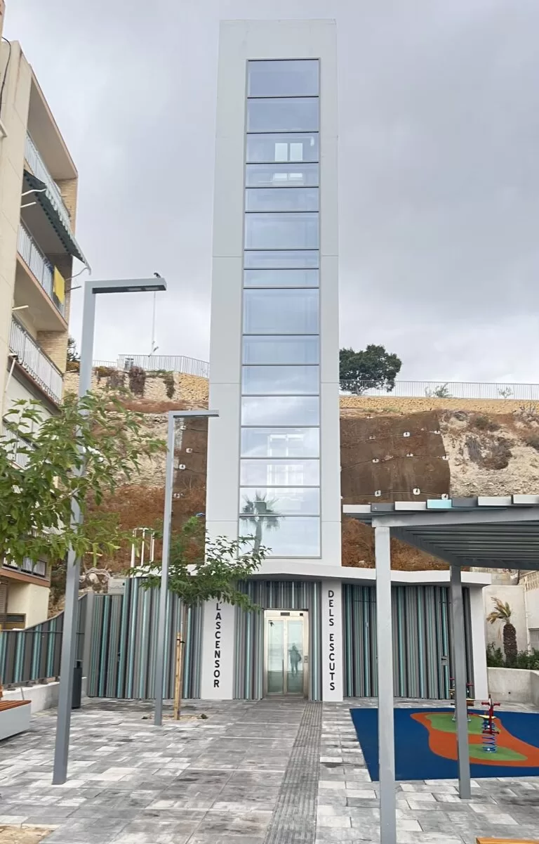 L'Ascensor dels Escuts in Villajoyosa, a glass public lift tower rising from a small square with a playground, linking the beach with the Barberes Sur district above
