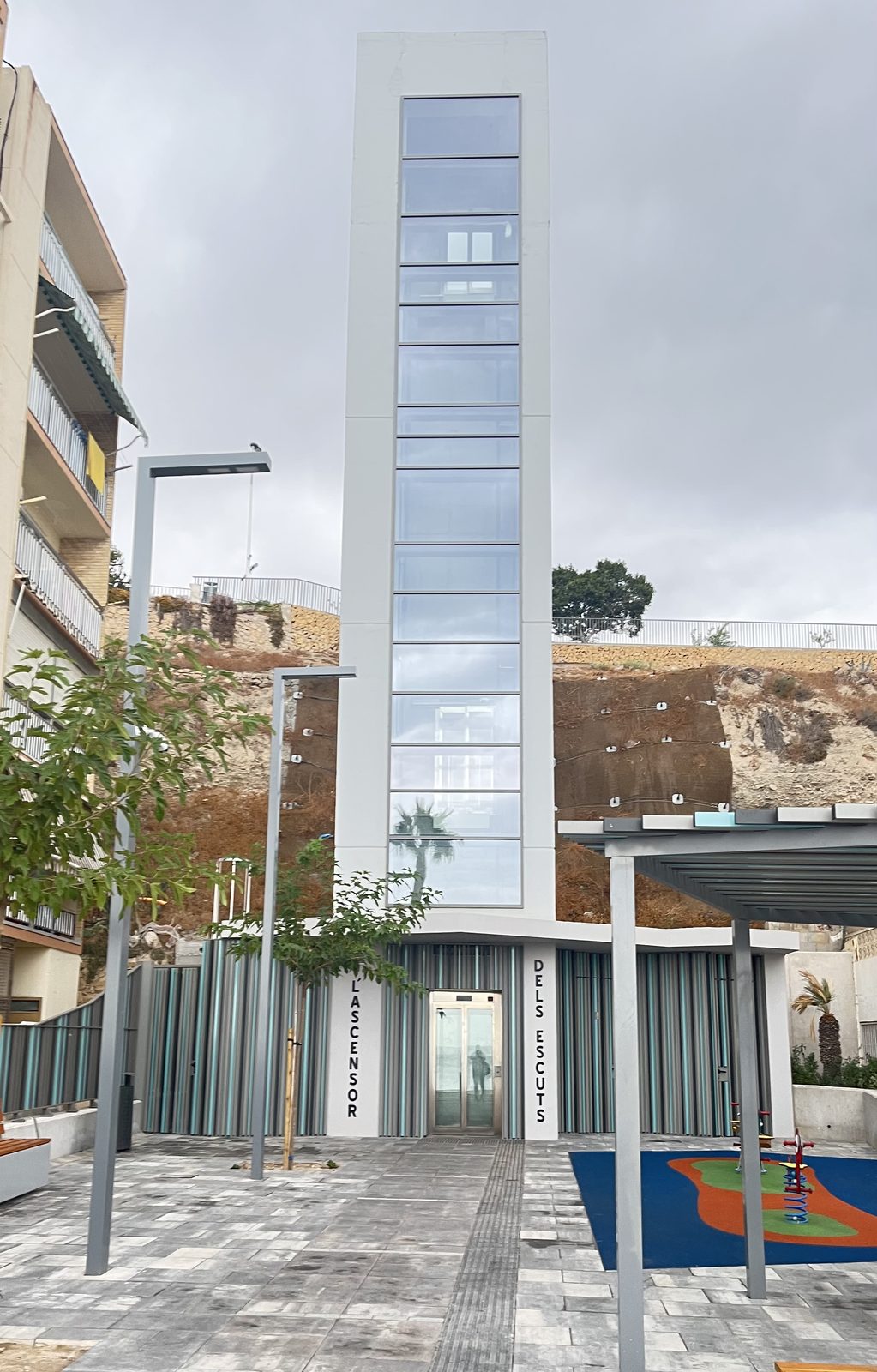 L'Ascensor dels Escuts in Villajoyosa, a glass public lift tower rising from a small square with a playground, linking the beach with the Barberes Sur district above